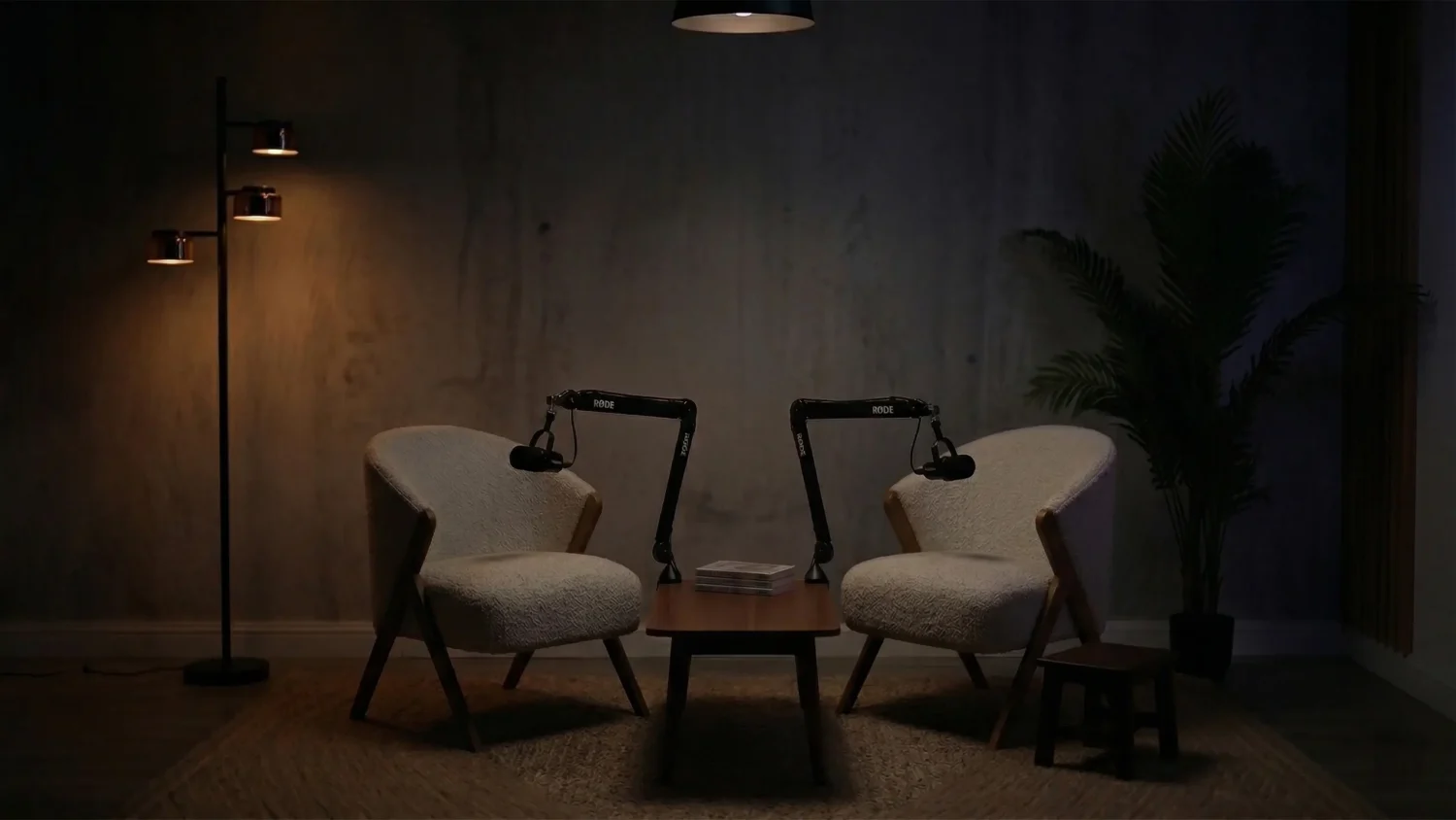 A moody, warmly lit podcast recording set featuring two white textured armchairs, professional microphones on black boom arms, and a minimalist floor lamp against a concrete wall.
